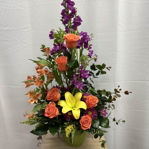 Tall mixed flower arrangement with orange roses, purple blooms, and a yellow lily in a vase