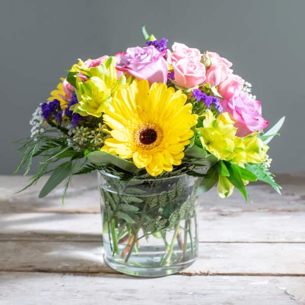 Mixed bouquet with yellow gerbera daisies, pink roses, and purple accents in a glass vase