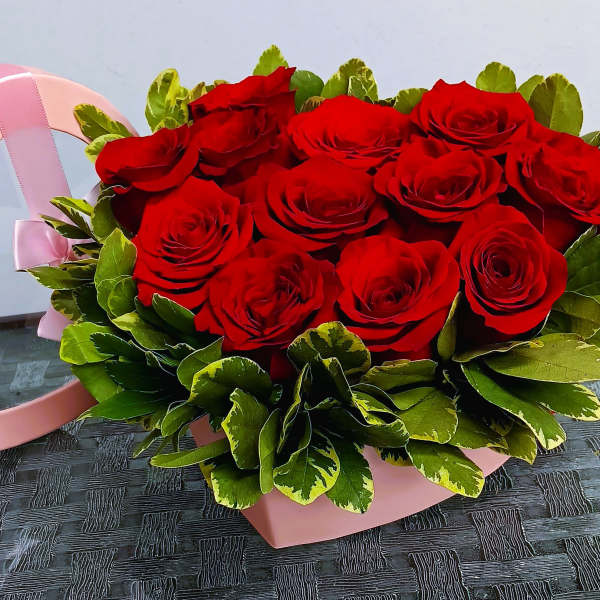 Red roses arranged in a pink heart-shaped box with green foliage