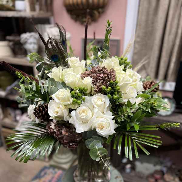 White roses arranged in a glass vase with peacock feathers and pinecones