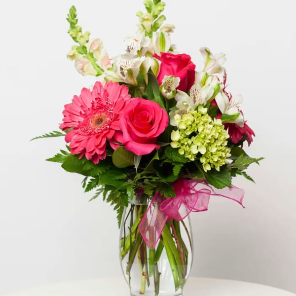 Pink gerbera daisies and roses in a clear glass vase with a pink ribbon