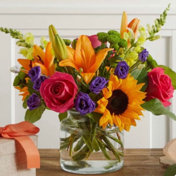 Bright mixed bouquet in a clear glass vase with roses, lilies, sunflowers, and purple blooms