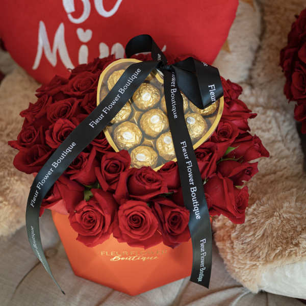Red roses arranged around a heart-shaped box of chocolates with a black ribbon.