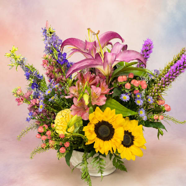 Mixed bouquet with purple lilies, sunflowers, and pink blooms in a white vase