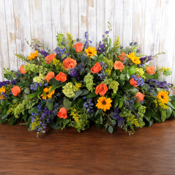 Low spray arrangement of orange roses, yellow daisies, and purple flowers in an elongated shape