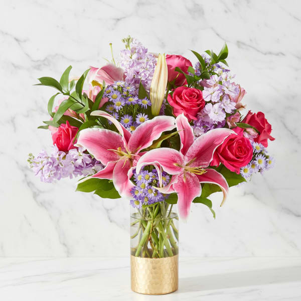 Pink lilies and roses arranged in a glass vase with purple filler flowers