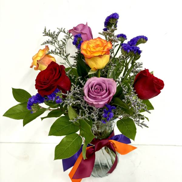 Mixed bouquet of red, yellow, and lavender roses with purple filler flowers in a glass vase with colorful ribbons