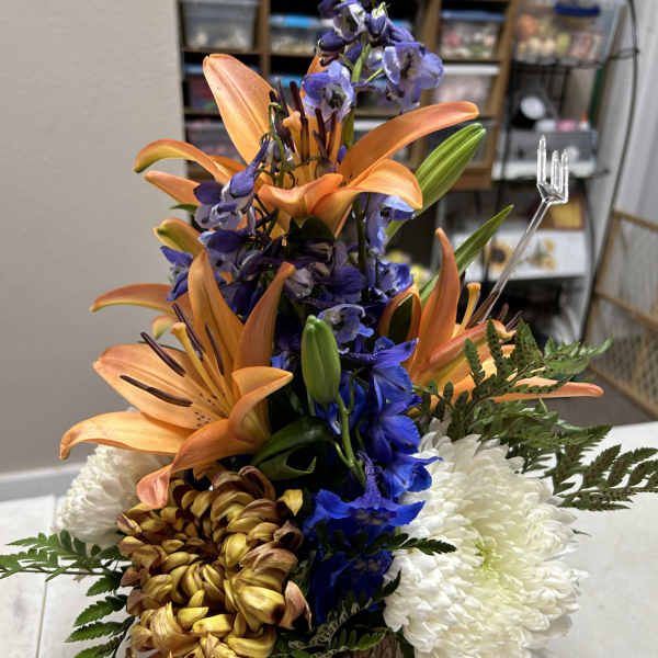 Orange lilies with blue delphinium and white chrysanthemums in a wood-look container