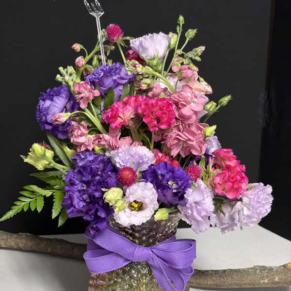 Colorful bouquet of pink and purple flowers in a textured glass vase with a purple ribbon