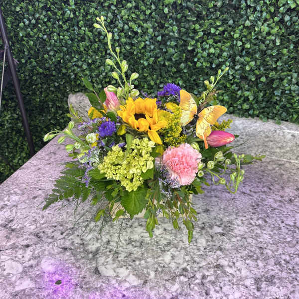 Colorful flower arrangement with a sunflower, pink blooms, and a butterfly pick in a low container