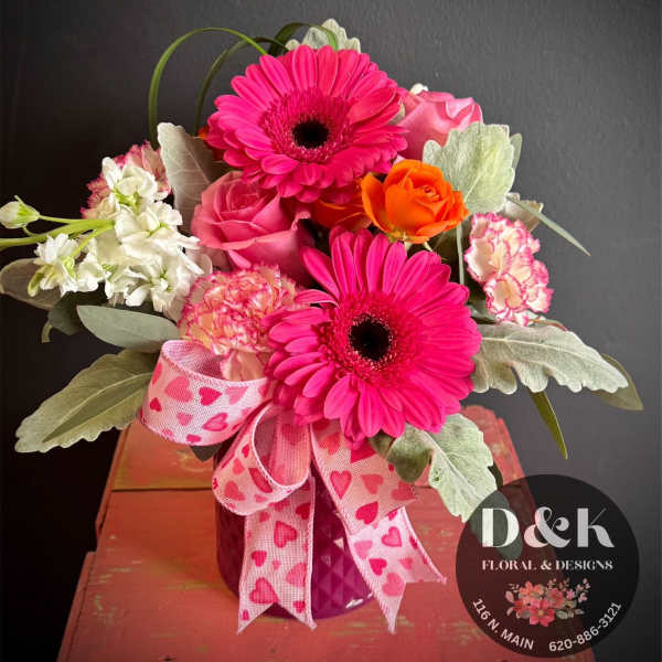 Pink gerbera daisies and roses in a purple vase with a heart ribbon