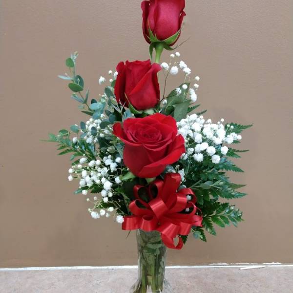 Three red roses in a clear glass vase with baby's breath and a red ribbon