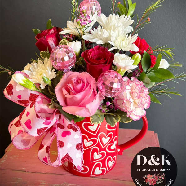 Pink and red flower arrangement in a heart-covered mug with a ribbon bow