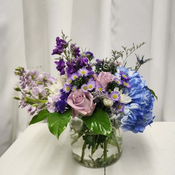 Mixed bouquet in a clear glass vase with blue hydrangea, pink roses, and purple flowers