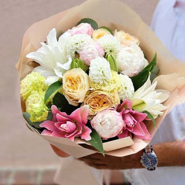 Hand-tied bouquet of white and pink lilies, roses, peonies, and mixed blooms wrapped in beige paper.