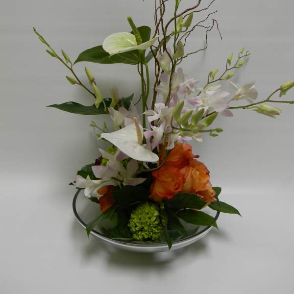 Tall floral arrangement with white orchids and orange roses in a shallow glass bowl