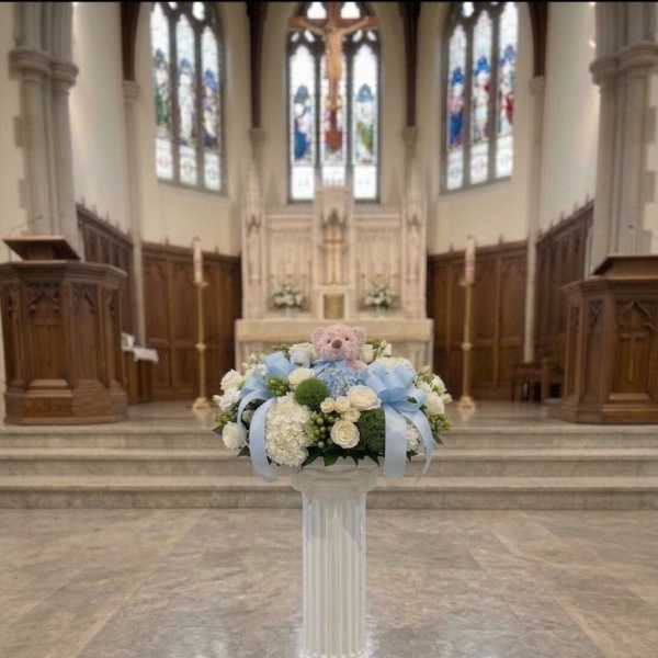 Pastel floral arrangement with a teddy bear on a white pedestal in a church