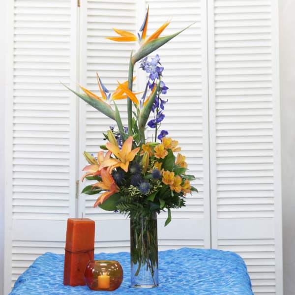 Tall tropical flower arrangement in a clear glass vase with orange and blue blooms