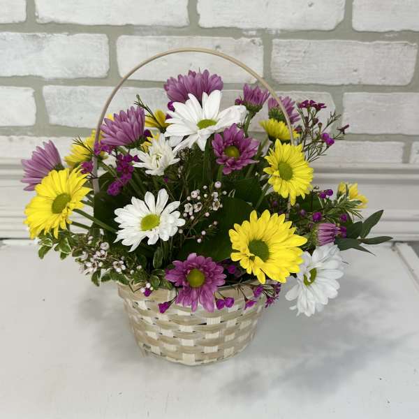 Basket arrangement of yellow, white, and purple daisies with small pink filler flowers.