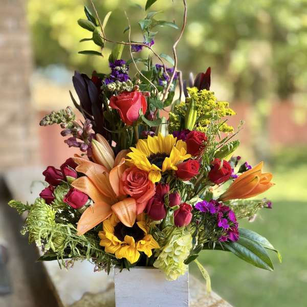 Mixed bouquet in a white square vase with roses, sunflowers, lilies, and purple blooms