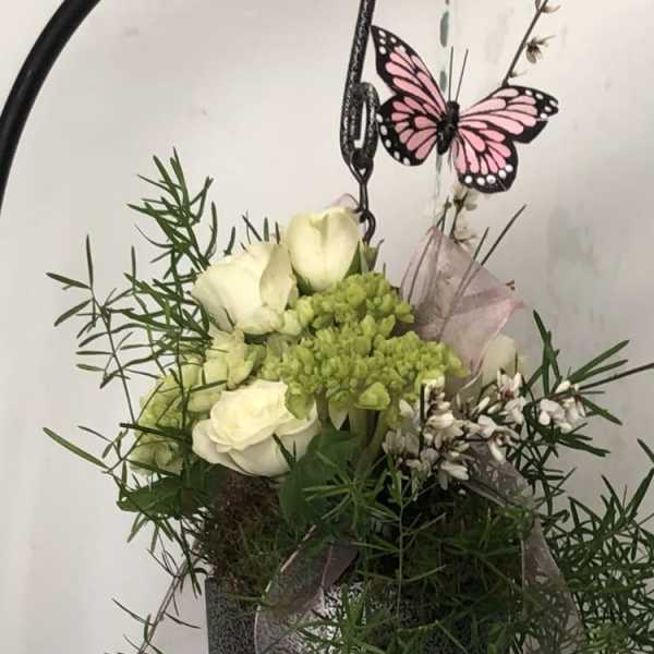 White roses and green blooms in a hanging metal basket with a pink butterfly decoration.