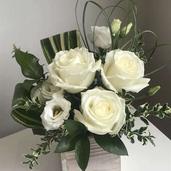 White roses and lisianthus in a square wooden vase with looping grass accents