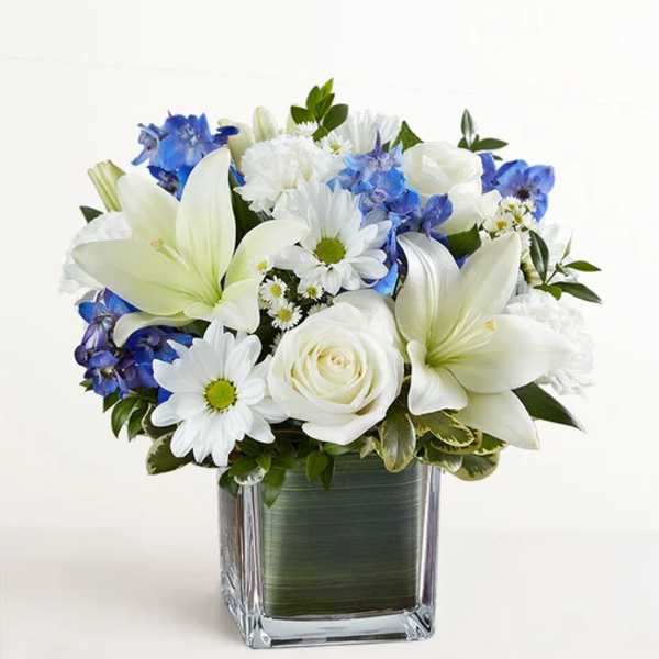 White lilies, roses, and blue flowers in a square glass vase