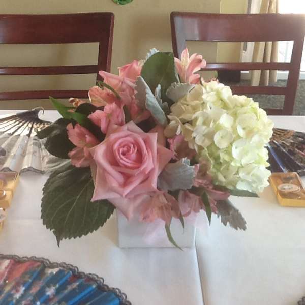 Event centerpiece with pink roses and white hydrangeas in a white cube vase on a table with fans.