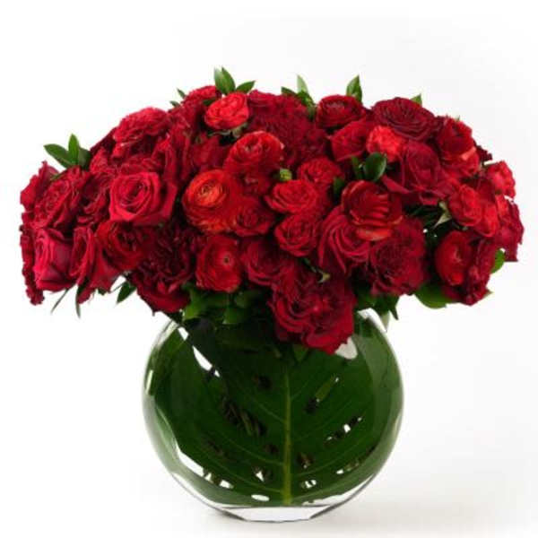 Low round arrangement of red roses and ranunculus in a clear glass vase lined with a large green leaf.