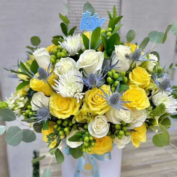 Hatbox arrangement of yellow and white roses with mums, blue thistles, and an It’s a Boy pick.