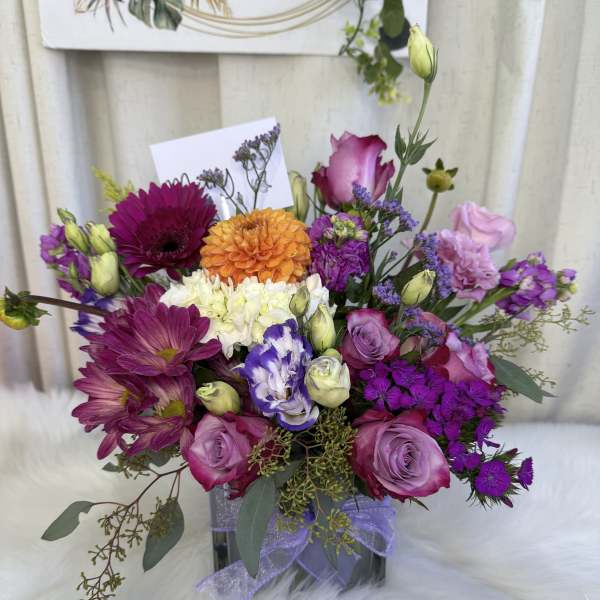 Mixed bouquet of purple, pink, orange, and white flowers in a square vase with ribbon.