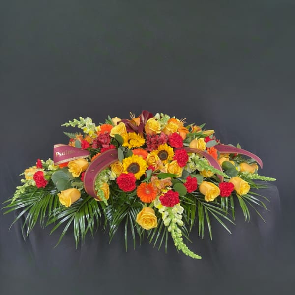 Elongated arrangement of yellow roses, orange gerberas, sunflowers, and red carnations with burgundy ribbons
