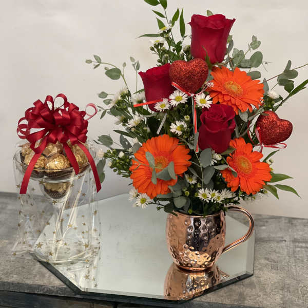 Red roses and orange gerbera daisies in a copper mug beside a gift jar of chocolates