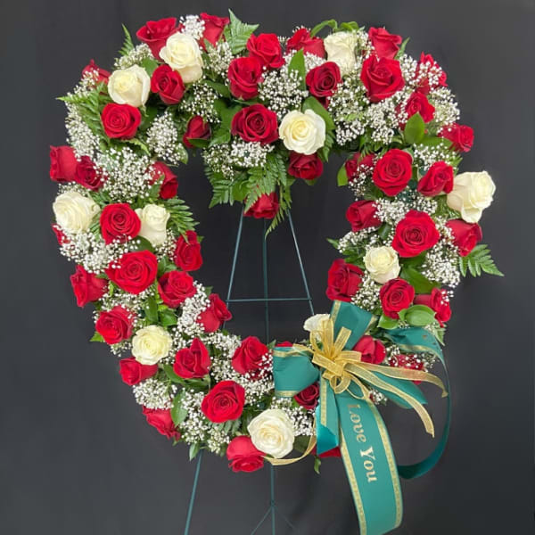 Heart-shaped standing wreath of red and white roses with white filler flowers and a green ribbon bow.