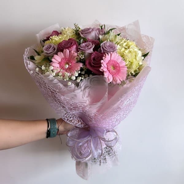 Handheld bouquet of pink gerberas, roses, and pale hydrangeas wrapped in lavender paper
