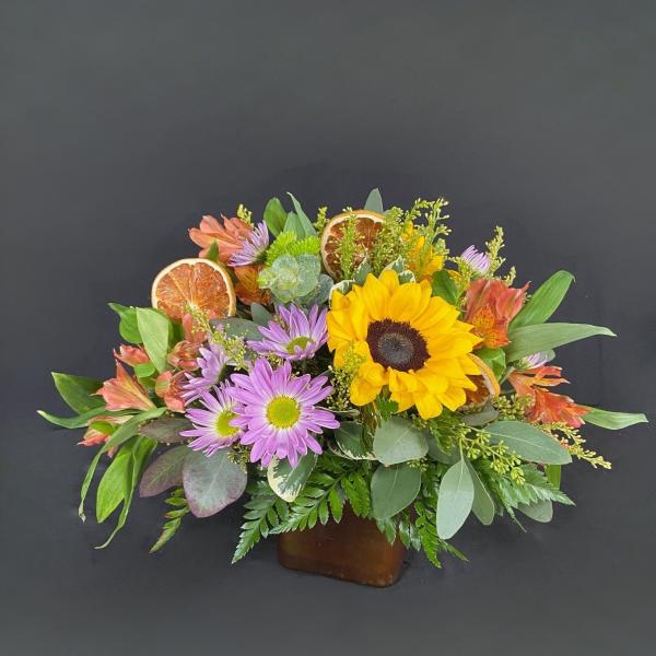 Mixed bouquet with a sunflower, purple daisies, and dried citrus slices in a low container