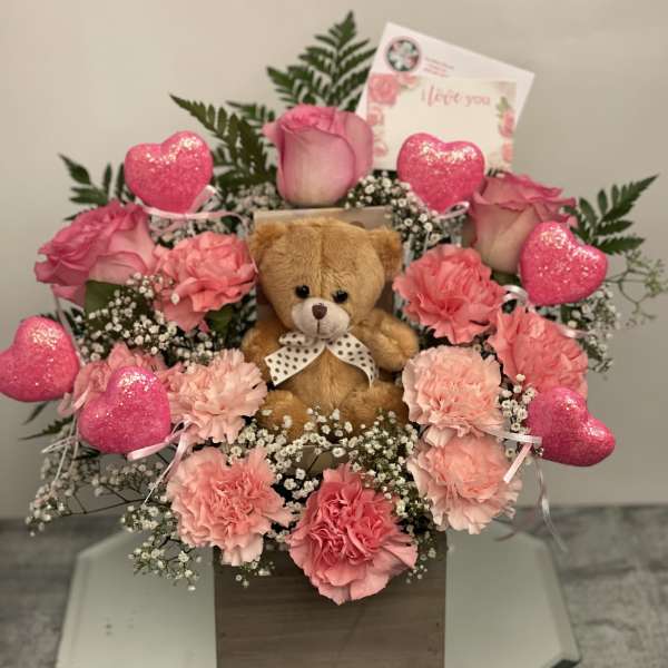 Pink carnations and roses arranged with a teddy bear and heart decorations