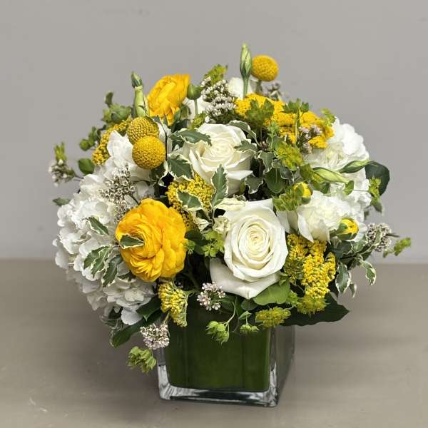 Rounded arrangement of white roses, hydrangeas, and yellow blooms in a clear square glass vase