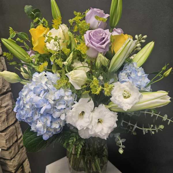 Mixed bouquet with blue hydrangeas, roses, lilies, and white blooms in a glass vase