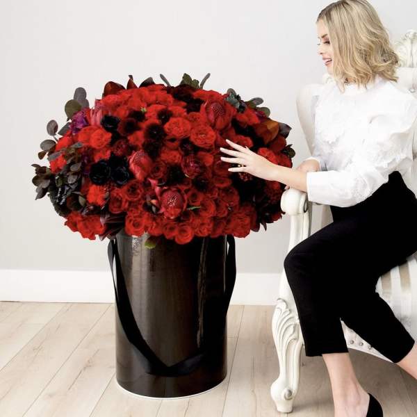 Large red rose arrangement in a tall black cylinder box beside a seated woman