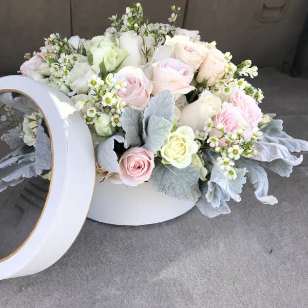 Pastel roses and small white flowers in a round white box with a clear lid.