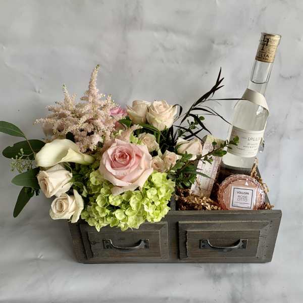 Pink and cream roses with hydrangea in a wooden drawer box beside a wine bottle and candle.