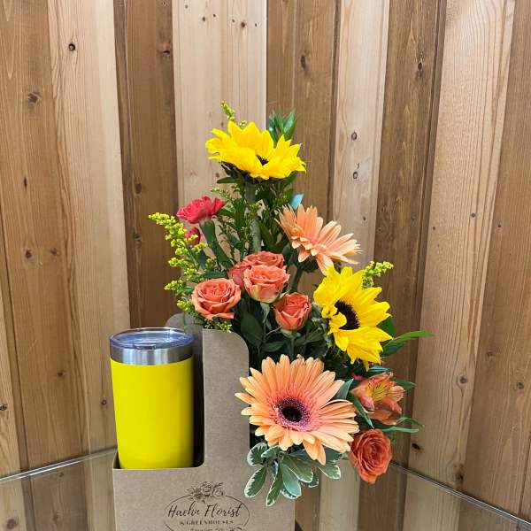 Mixed bouquet with sunflowers, roses, and gerbera daisies in a gift box beside a yellow tumbler