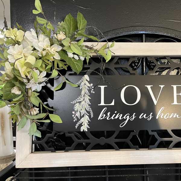 Framed sign with white floral accents and the words "LOVE brings us home"