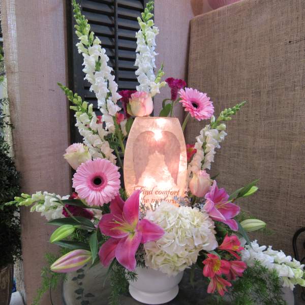 Pink lilies and gerbera daisies arranged around a glowing memorial candle.