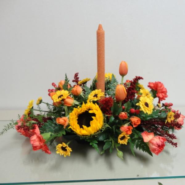 Low floral centerpiece with a tall orange candle and mixed orange, yellow, and red blooms