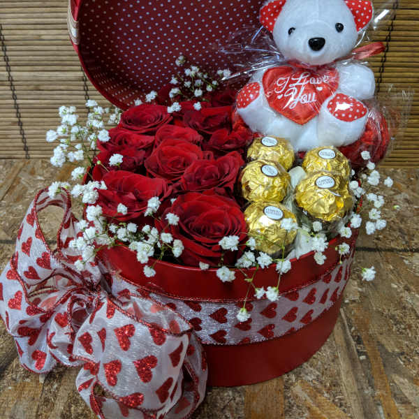 Red hat box filled with red roses, gold-wrapped chocolates, and a white teddy bear holding a heart.
