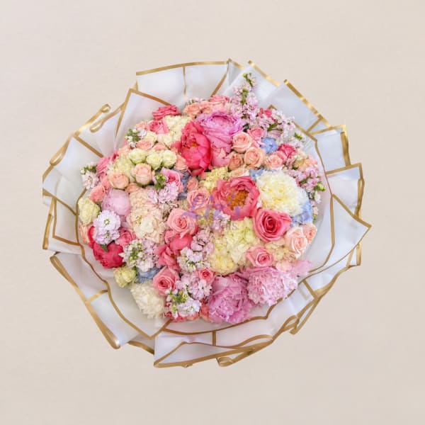 Round bouquet of pink, peach, and white flowers wrapped in white paper with gold trim.