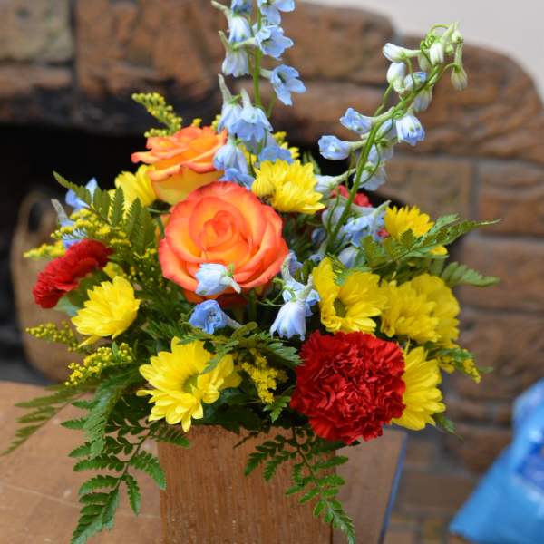 Colorful flower arrangement in a wooden cube vase with roses, daisies, and blue delphinium