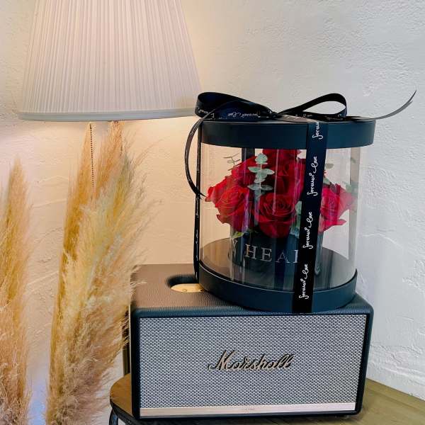 Red roses in a round gift box on a speaker, with pampas grass nearby.